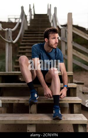 Ginnastica maschile con bearded in abiti sportivi seduti su scale di legno mentre si prepara ad allenarsi e guardare via Foto Stock