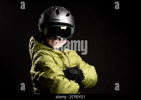 Piccolo capretto sportivo messo a fuoco in vestiti invernali con goggle di sci su sfondo nero in studio guardando la fotocamera Foto Stock