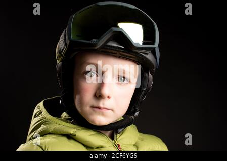 Piccolo capretto sportivo messo a fuoco in vestiti invernali con goggle di sci su sfondo nero in studio guardando la fotocamera Foto Stock