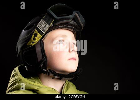 Piccolo capretto sportivo messo a fuoco in vestiti invernali con goggle di sci su sfondo nero in studio che guarda via Foto Stock