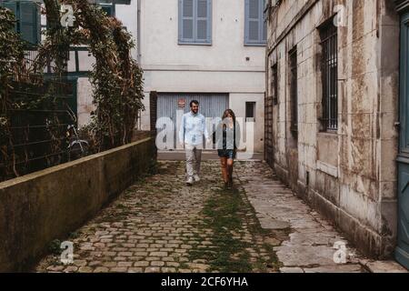 Giovane coppia allegra in abiti casual di stile che tengono le mani e. sorridente mentre si cammina lungo la vecchia strada stretta della città Foto Stock