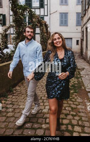 Giovane coppia allegra in abiti casual di stile che tengono le mani e. sorridente mentre si cammina lungo la vecchia strada stretta della città Foto Stock