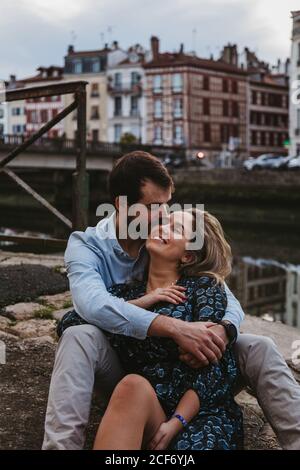 Giovane coppia positiva in abiti casual godendo di una data romantica mentre siediti insieme al confine in pietra della città con edifici antichi in background Foto Stock