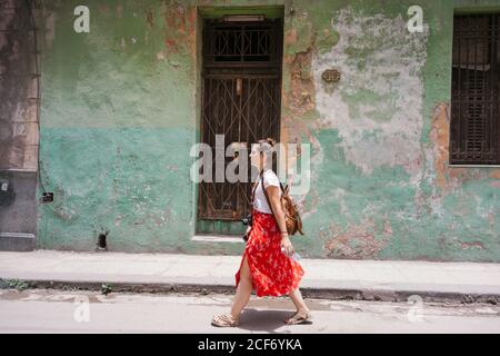 Vista laterale della donna in vacanza in gonna rossa colorata con zaino e fotocamera a piedi lungo la strada con la vecchia Edificio sullo sfondo a Cuba Foto Stock