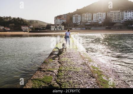Giovane donna che cammina con il cane sul molo di pietra a Lekeitio Città nei Paesi Baschi con edifici residenziali e colline in sfondo Foto Stock