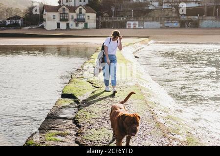 Giovane donna che cammina con il cane sul molo di pietra a Lekeitio Città nei Paesi Baschi con edifici residenziali e colline in sfondo Foto Stock