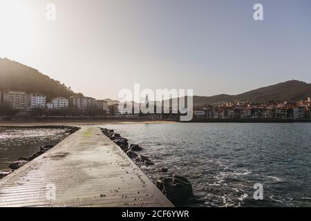 Incredibile scenario con molo in legno bagnato che conduce tra massi e. piccole onde marine verso il lungomare della città turistica a piedi Di colline boscose sotto il cielo senza nuvole in Spagna Foto Stock