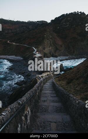 Pietra di pavimentazione vuota che conduce lungo il ponte tra recinzioni di pietra e. lato di collina rocciosa lungo la riva di lavaggio da mare tormentato Acqua con onde di schiuma bianca al crepuscolo in Spagna Foto Stock