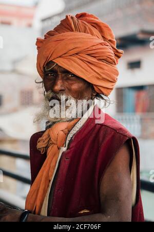 Varanasi, India - FEBBRAIO, 2018: Uomo indù invecchiato in tartarughe arancioni con giubbotto rosso senza maniche in piedi sulla strada e guardando la macchina fotografica Foto Stock