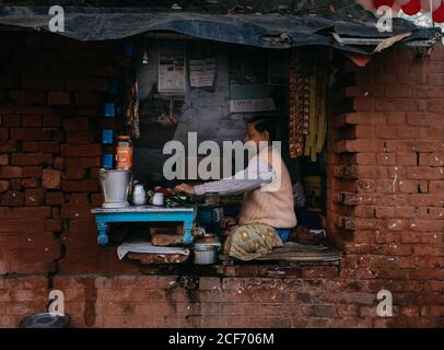 Varanasi, India - FEBBRAIO, 2018: Vista laterale di maschio adulto in abiti tipici preparare il cibo mentre si siede con le gambe incrociate sul buco della finestra di vecchio edificio in mattoni Foto Stock