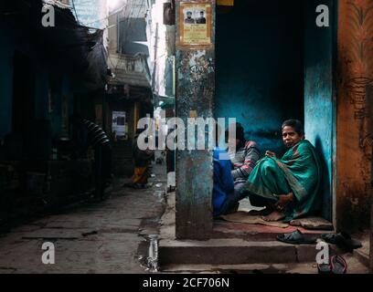 Varanasi, India - Febbraio, 2018: Vista laterale di una povera donna anziana che guarda la macchina fotografica mentre siede con altre persone senza tetto vicino al vecchio edificio dilapidata sulla stretta strada della città Foto Stock