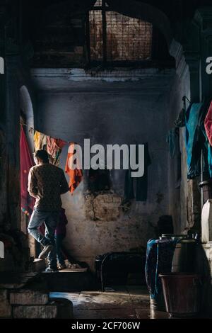 Varanasi, India - Febbraio, 2018: Vista posteriore del povero uomo con il capretto in piedi nella porta con luce dalla strada mentre lascia la casa miserabile con pochi utensili e vestiti appesi sulla corda Foto Stock