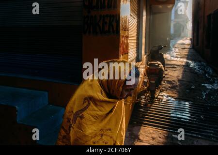 Varanasi, India - Febbraio, 2018: Vista laterale della vecchia femmina indiana in shawl tradizionale zafferano colorato in piedi accanto alla panetteria locale su strada stretta sporca Foto Stock
