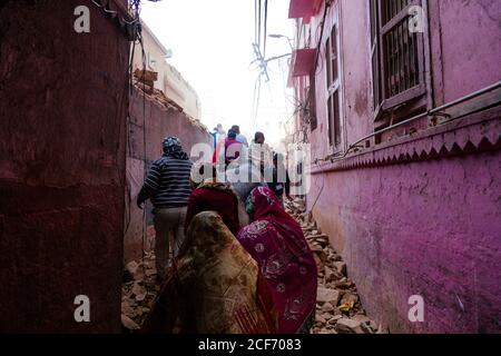 Varanasi, India - Febbraio, 2018: Vista posteriore della gente locale e mucca che cammina su una strada stretta disseminata di pietre da un edificio in rovina Foto Stock
