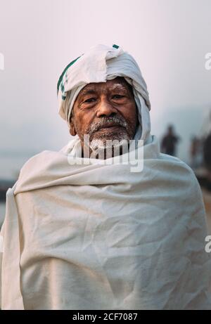 Varanasi, India - FEBBRAIO, 2018: Primo piano ritratto di un anziano indiano che indossa abiti bianchi tradizionali in piedi sulla riva del fiume Foto Stock