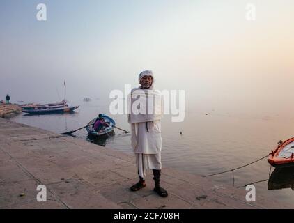 Varanasi, India - FEBBRAIO, 2018: Anziano uomo indiano che indossa abiti bianchi tradizionali in piedi sulla riva del fiume con barche da pesca in acqua nella mattina presto foggy Foto Stock