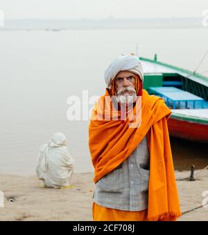 Varanasi, India - FEBBRAIO, 2018: Anziano uomo indiano che indossa abiti tradizionali arancioni in piedi sulla riva del fiume con barche da pesca in acqua nella mattina presto foggy Foto Stock