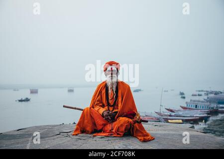 Varanasi, India - FEBBRAIO, 2018: Uomo indù alato in turbano arancione seduto sulla strada e guardando la macchina fotografica Foto Stock
