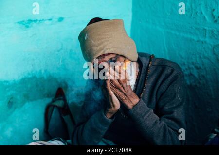 Varanasi, India - Febbraio, 2018: Povero anziano uomo indù in berretto lavorato beige seduto nell'angolo della casa con pareti blu e sigaro fumante Foto Stock