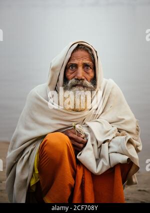 Varanasi, India - FEBBRAIO, 2018: Anziano uomo indiano che indossa abiti tradizionali arancioni seduti sulla riva del fiume con barche da pesca in acqua nella mattina presto foggy Foto Stock