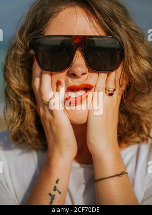 Elegante donna dai capelli ricci marroni con rossetto rosso in occhiali da sole alla moda guardando la macchina fotografica mentre si stringe il viso nei palmi Foto Stock