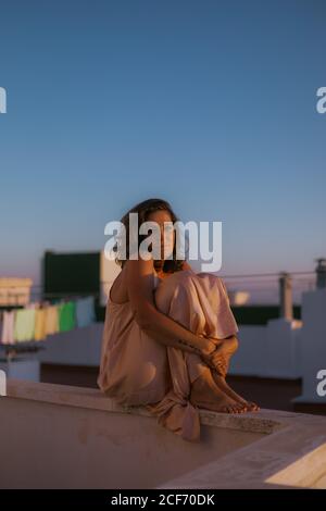 Giovane donna dai capelli ricci in elegante abito di seta rosa seduto su recinto di balcone con le braccia intorno alle gambe e guardando la macchina fotografica durante il tramonto estivo Foto Stock