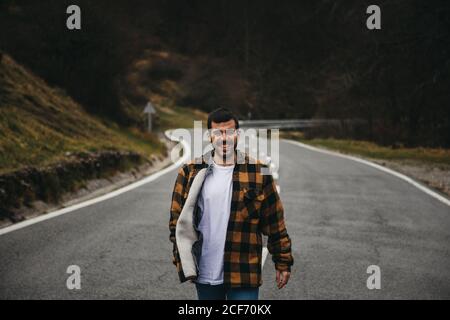 Uomo allegro con capelli neri e bearded in abbigliamento casual sorridente e. guardando la fotocamera mentre si è in piedi su una strada vuota in autunno campagna Foto Stock