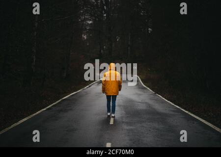 Vista posteriore di un uomo anonimo in giacca gialla che cammina strada asfaltata vuota tra foresta verde Foto Stock