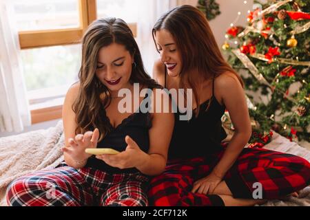 Due amici donne che si divertono nelle vacanze invernali a casa e che guardano qualcosa sul cellulare vicino albero di natale in interni accoglienti. Interni con decorazioni natalizie. Foto Stock