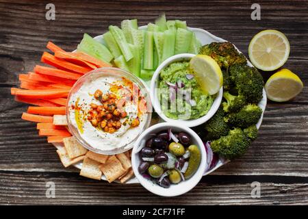 Dall'alto appetitoso hummus sano salsa verde cut cetrioli di carote e i cetrioli salati di broccoli verdi decorati con limone a fette ciotole bianche su tavola di legno Foto Stock