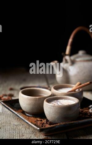 Masala chai servito in ciotole di ceramica con anice stellato e. bastoncini di cannella disposti su un tavolo di legno con teiera e pezzo di stoffa Foto Stock