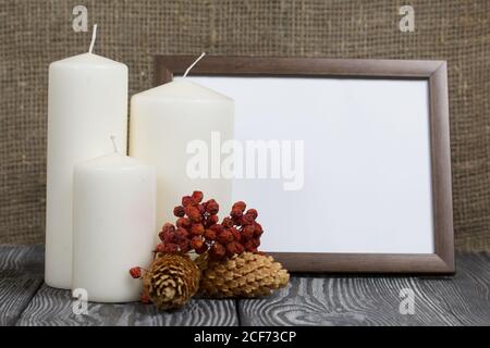 Cornice fotografica in legno con un campo bianco per un'iscrizione o un'immagine. Nelle vicinanze si trovano candele con coni di abete e bacche di rowan essiccate. Foto Stock