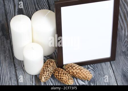 Cornice fotografica in legno con un campo bianco per un'iscrizione o un'immagine. Nelle vicinanze si trovano candele con coni di abete. Foto Stock