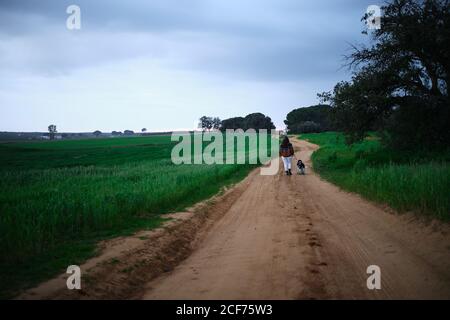 Vista posteriore della donna senza volto in vestito casual a piedi con il cane su vuota strada rurale tra campi verdi in campagna Foto Stock