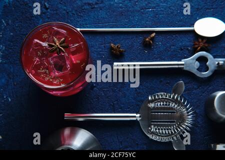 Vista dall'alto di una gustosa bevanda rossa con cubetti di ghiaccio e. set di attrezzature per barman su tavolo blu Foto Stock