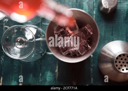 Da sopra il processo di versamento della bevanda alcolica rossa dal flacone in shaker con cubetti di ghiaccio durante la preparazione del cocktail al bancone Foto Stock