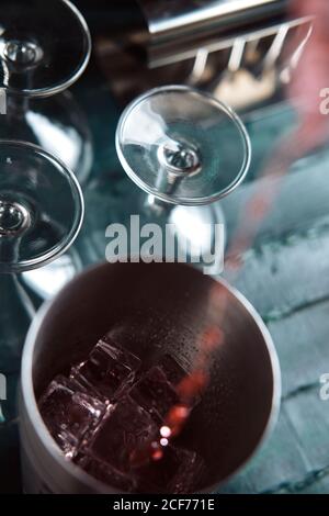 Da sopra il processo di versamento della bevanda alcolica rossa dal flacone in shaker con cubetti di ghiaccio durante la preparazione del cocktail al bancone Foto Stock