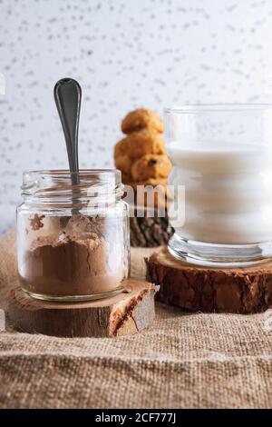 Vasetto trasparente di cacao in polvere e bicchiere di latticini in composizione con gustosi biscotti marroni sfocati su coste di legno in piedi su un panno contro una parete bianca macchiata Foto Stock
