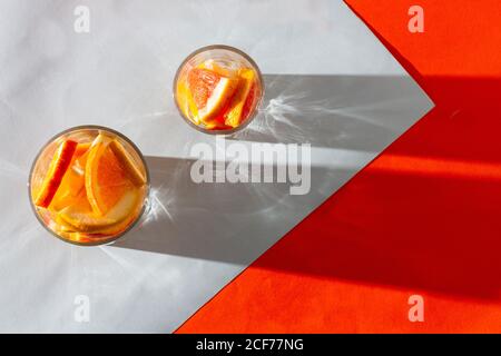 Dall'alto fette fresche di arancio e limone in tazze di vetro proietta l'ombra su sfondo bianco arancione Foto Stock