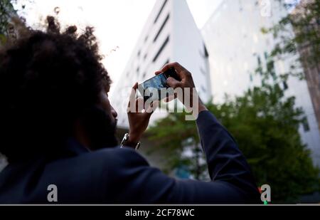 Vista posteriore di imprenditore etnico maschile di tendenza in abbigliamento formale scattare foto di un edificio residenziale bianco sfocato sul telefono cellulare Foto Stock