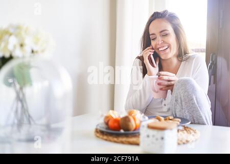 Attraente giovane donna felice utilizzando il telefono cellulare e facendo colazione al tavolo vicino alla finestra a casa Foto Stock
