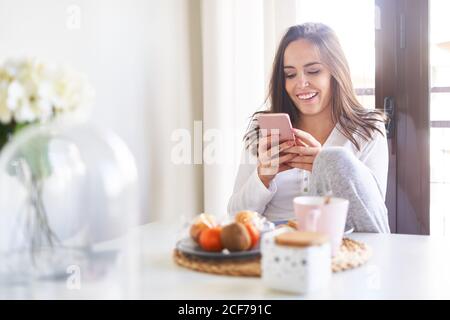 Attraente giovane donna felice utilizzando il telefono cellulare e facendo colazione al tavolo vicino alla finestra a casa Foto Stock