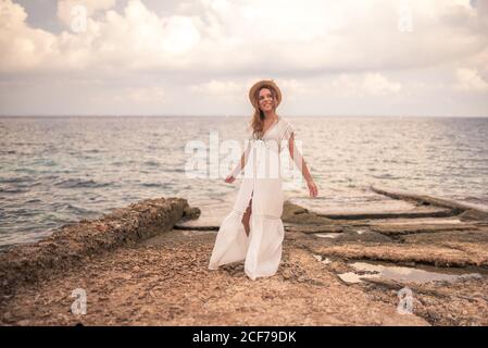 Carefree viaggiatore femminile in abito estivo e cappello in piedi pietroso mare sullo sfondo di mare stupefacente durante le vacanze Foto Stock