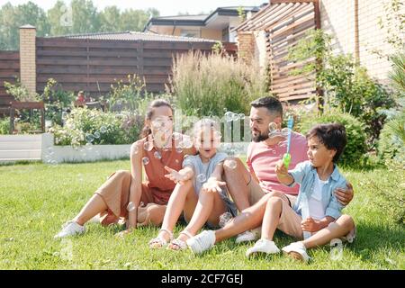 Famiglia moderna con due bambini che trascorrono la giornata estiva in cortile, ragazzino che fa bolle di sapone Foto Stock