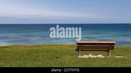 Panca di legno vuota in piedi vicino al bordo della riva di blu e tranquilla superficie del mare Foto Stock