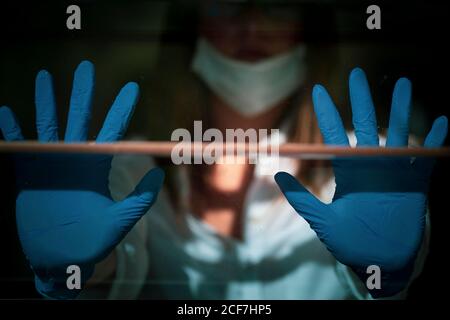 Donna giovane seria in maschera medica e guanti protettivi in piedi vicino alla finestra e guardando via durante l'isolamento e la quarantena a causa della pandemia del coronavirus Foto Stock