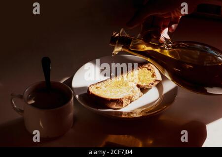 Da sopra raccolto persona anonima versando l'olio d'oliva dalla bottiglia su pane toast messo sul tavolo vicino tazza di caldo bere mentre si prepara la colazione a casa Foto Stock
