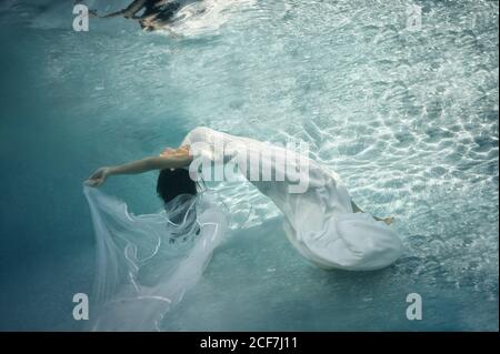 Bella bruna con capelli lunghi in lungo abito da sposa bianco con velare galleggiando sott'acqua con le braccia distese Foto Stock