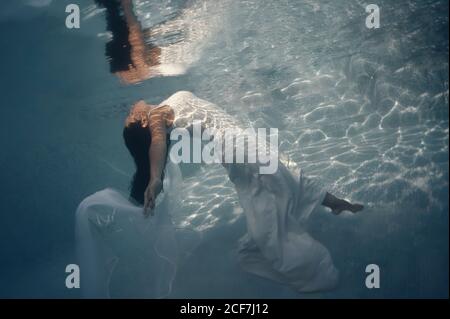 Vista laterale della bella bruna dai capelli lunghi in bianco lungo abito da sposa con velo galleggiante sott'acqua con braccia allungate Foto Stock