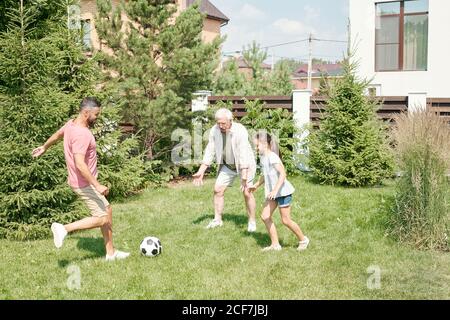 Giovane adulto che gioca a calcio con il padre anziano e. piccola figlia sul prato in cortile Foto Stock